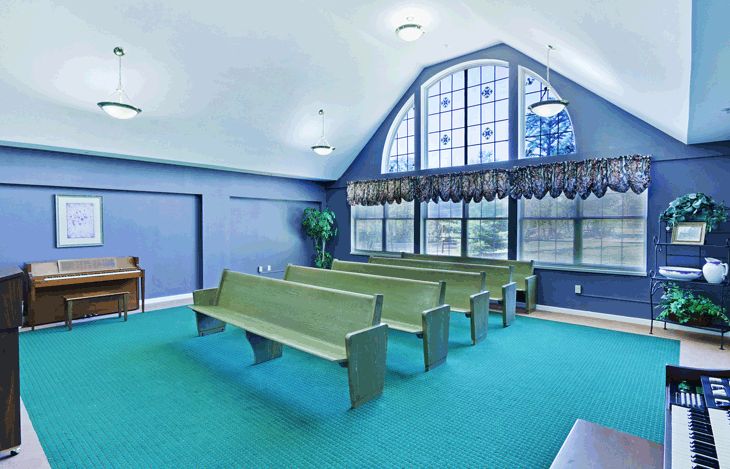 A chapel interior with green pews, large windows, and teal carpet.