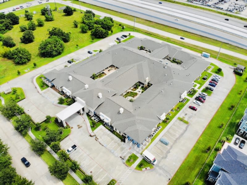 Aerial view of a large building surrounded by a parking lot and greenery, located near a busy road.