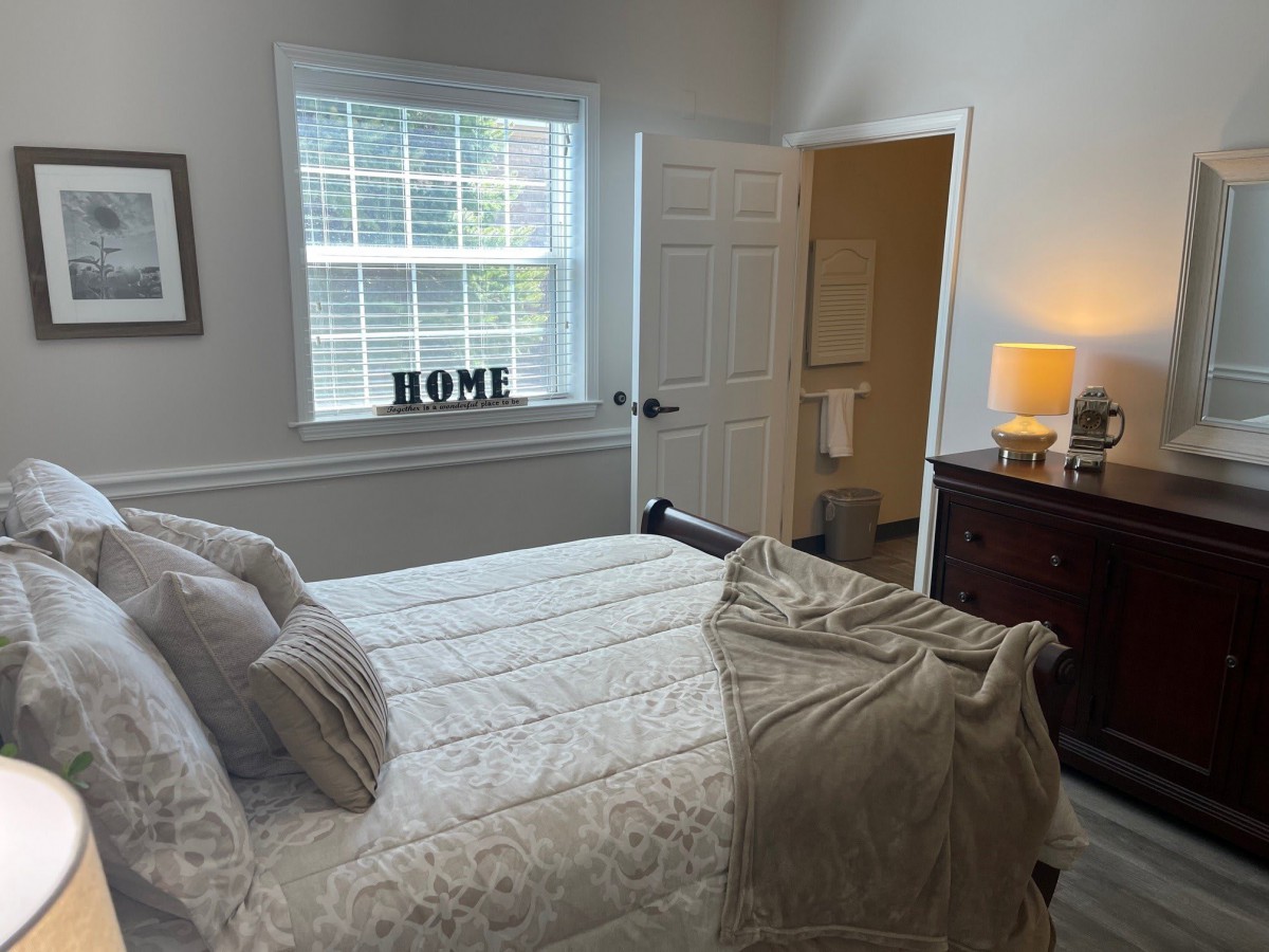 A cozy bedroom with a neatly made bed, a window with blinds, and soft lighting from a bedside lamp.