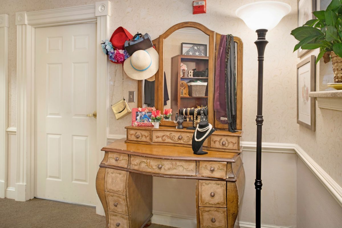 A vintage vanity dresser adorned with hats, scarves, and jewelry, accompanied by a floor lamp.