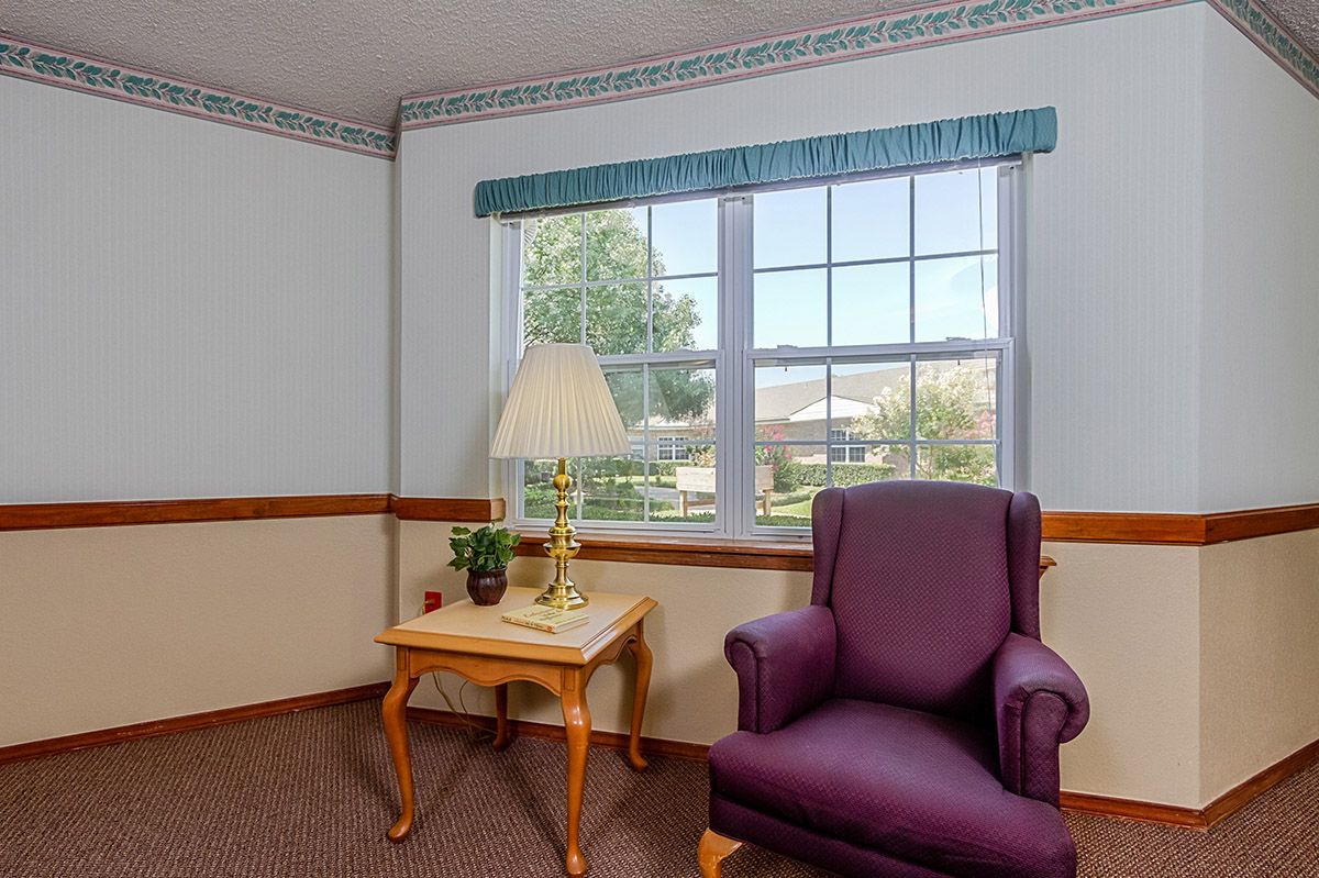 A cozy room corner with a purple armchair, side table with a lamp and plant, and a window view.