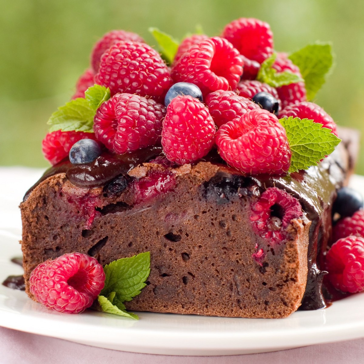A rich chocolate cake topped with fresh raspberries, blueberries, and mint leaves on a white plate.
