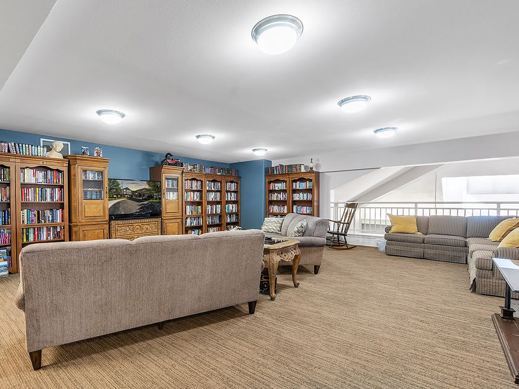 A spacious living room with comfortable sofas and large bookshelves, featuring warm lighting and a carpeted floor.
