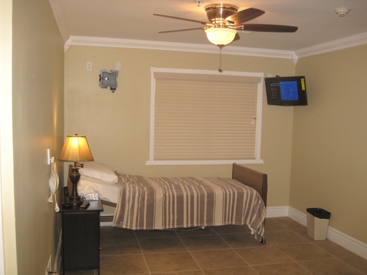 A small bedroom with a single bed, ceiling fan, wall-mounted television, and a lamp on a nightstand.