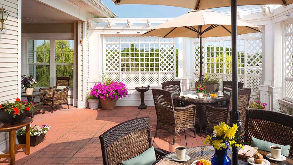 A sunny patio area with wicker chairs and a dining table, adorned with flowers and an umbrella.