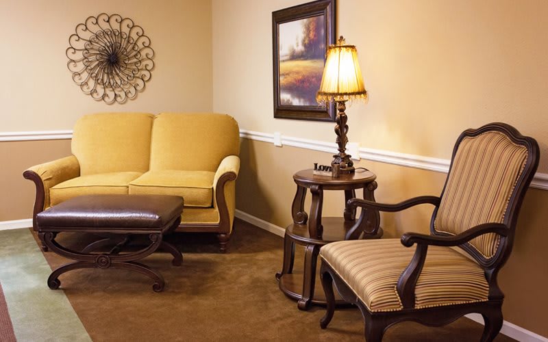 A cozy living room featuring a yellow sofa, an ornate lamp on a round wooden table, a striped chair, and artwork on the wall.