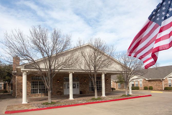 A brick building with a white porch and columns, accompanied by an American flag in the foreground.