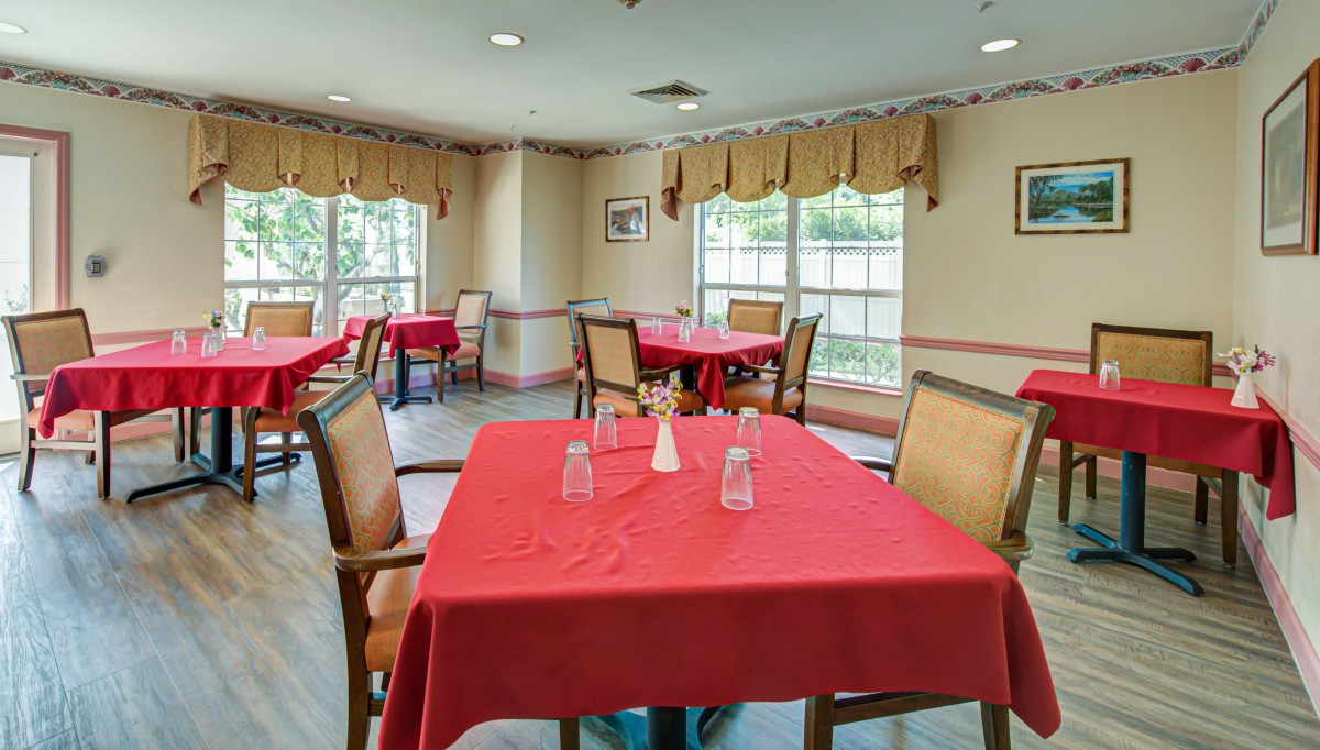 A dining room with tables covered in red tablecloths and chairs neatly arranged by the windows.