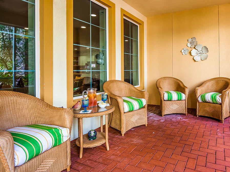 A cozy patio with wicker chairs featuring green and white striped cushions, a small table with drinks, and large windows.