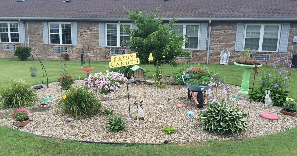 A charming garden with a variety of plants and flowers, featuring a sign and wheelbarrow in front of a house.