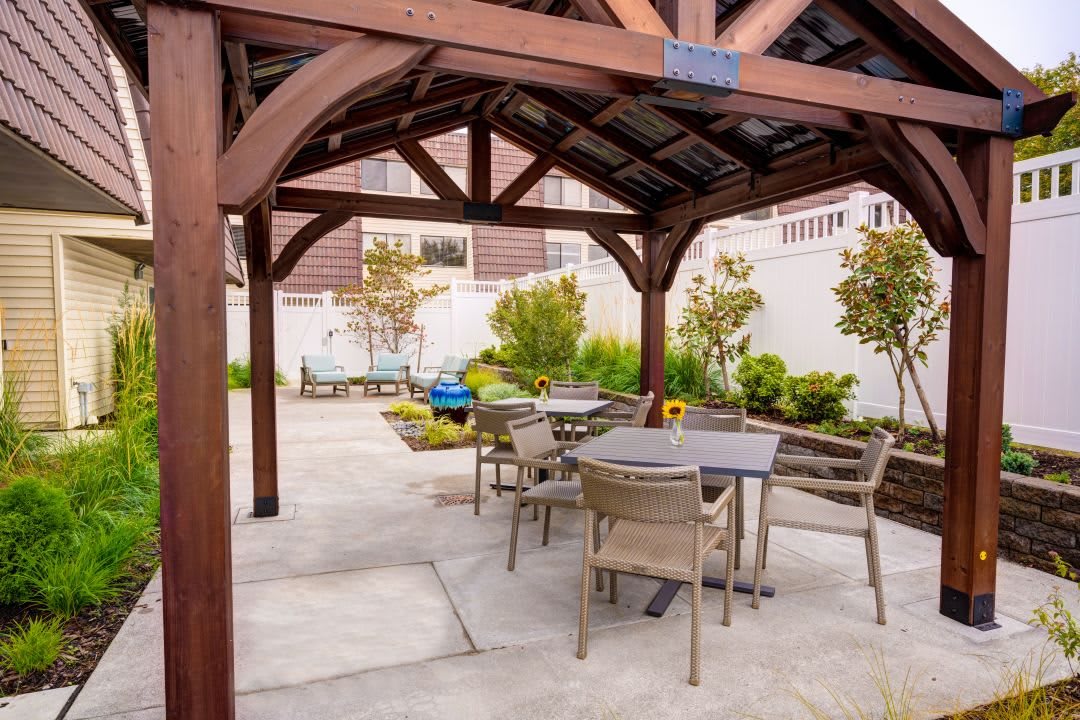 A cozy outdoor patio area with tables and chairs under a wooden gazebo, surrounded by a well-maintained garden.