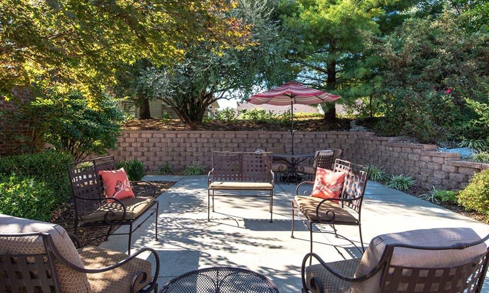 A cozy outdoor patio area with metal furniture and a garden umbrella, surrounded by trees and a brick wall.