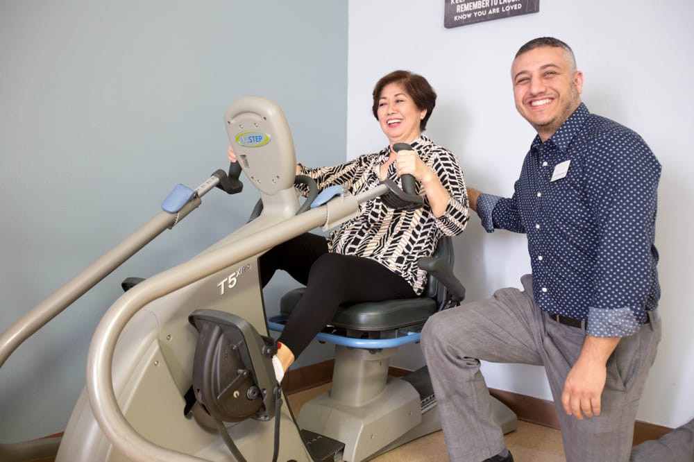A woman smiles while using a rehabilitation exercise machine, accompanied by a supportive individual.