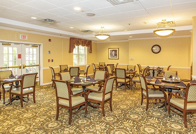 A spacious, well-lit dining room with neatly arranged tables and chairs, featuring an ornate carpet design.