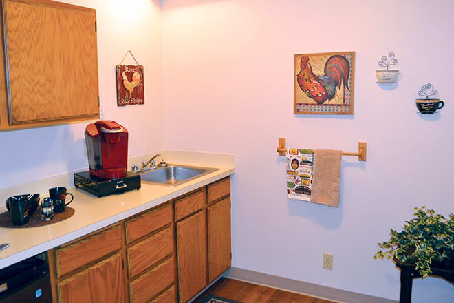 A small kitchen area featuring a coffee maker and wood cabinets, adorned with wall art and a towel rack.