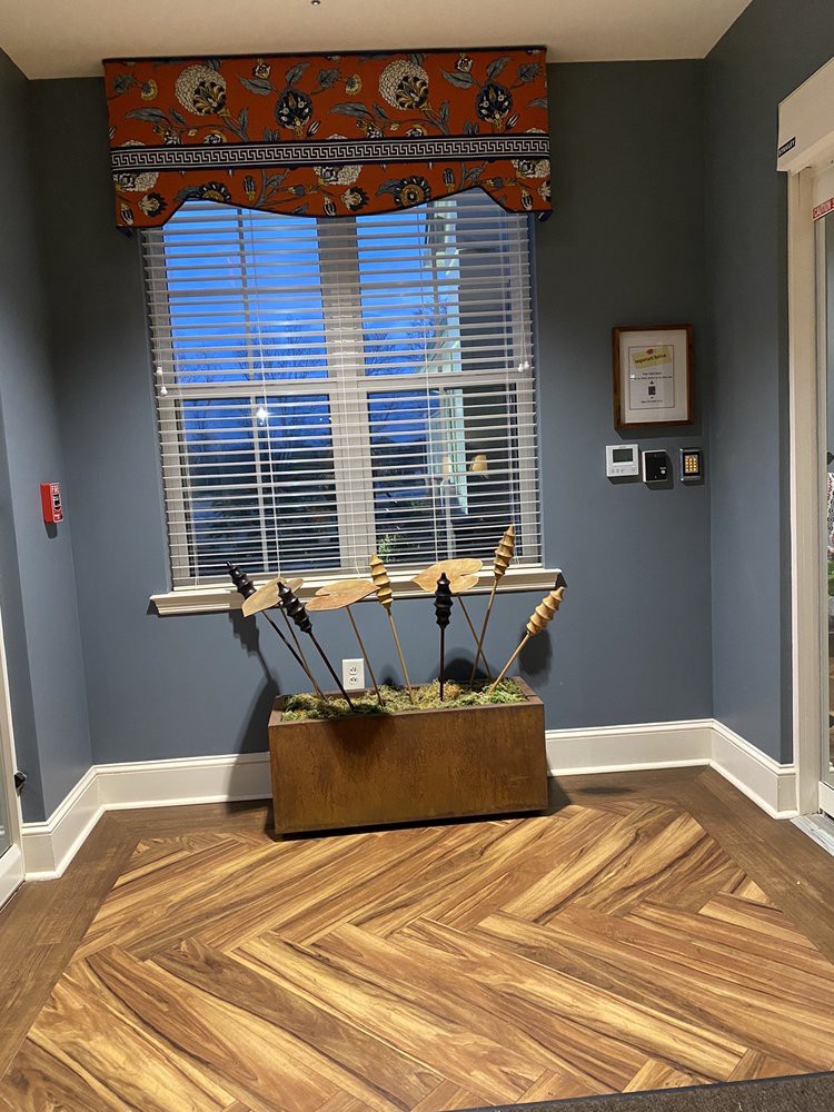 A cozy interior space featuring a window with blinds, a wooden floor, and decorative elements including a planter and artwork.