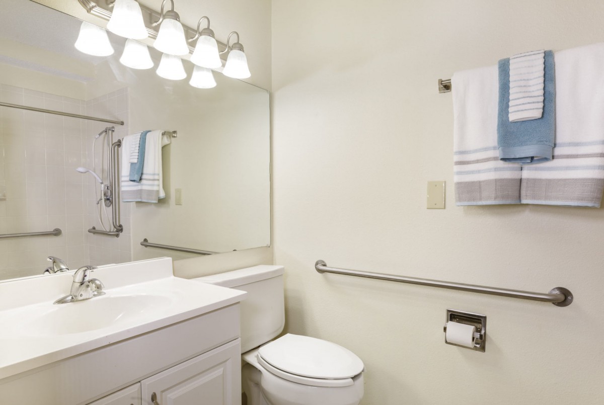 A clean and bright bathroom with a toilet, sink, and mirror, featuring a towel rack and overhead lighting.