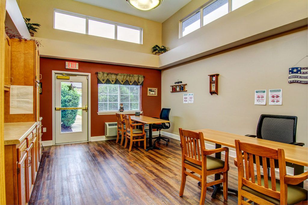 A cozy dining area with wooden floors, tables, and chairs, featuring large windows for natural light.