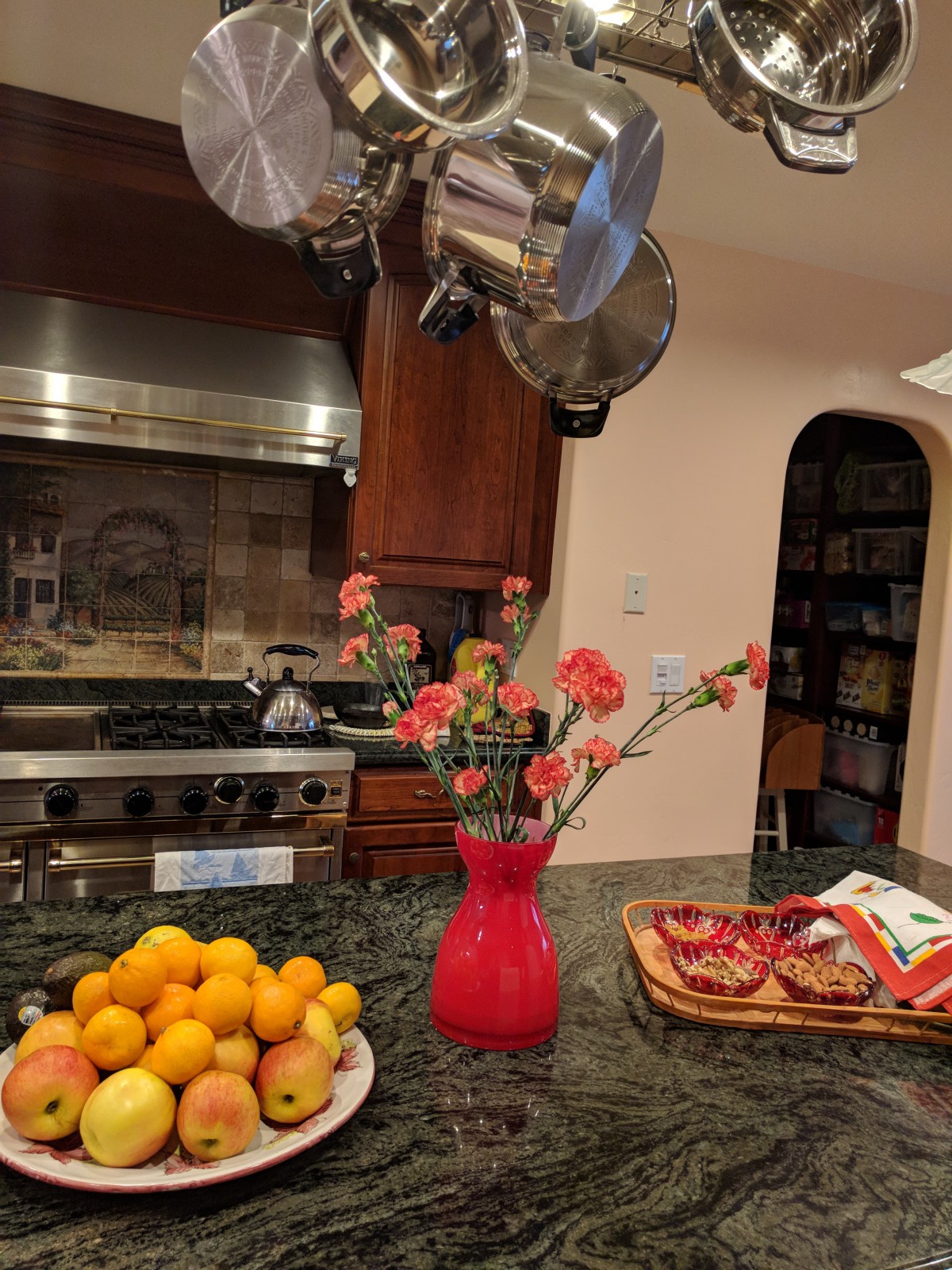 A kitchen with a granite countertop displaying a bowl of oranges, a red vase with flowers, and snacks in a basket.