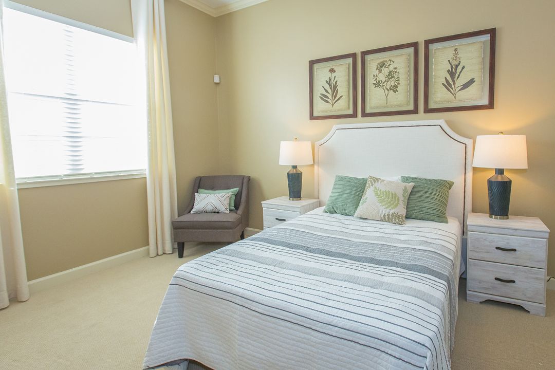 A cozy bedroom featuring a neatly made bed with striped bedding, decorative pillows, and three framed artworks above the headboard. A comfortable chair sits in the corner near a window, with a lamp on the bedside table completing the room's warm ambiance.