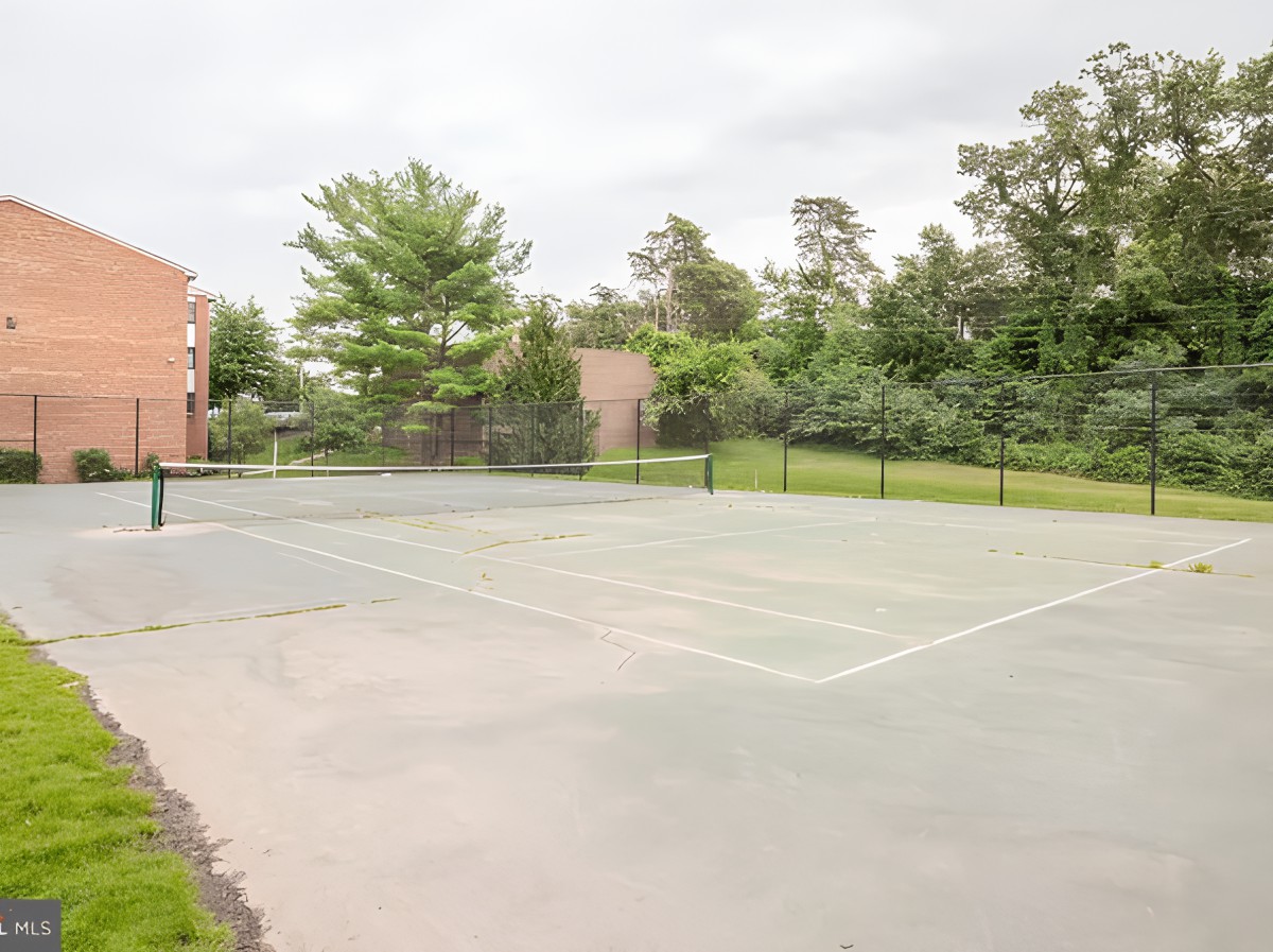 An empty outdoor tennis court surrounded by trees and a fence.
