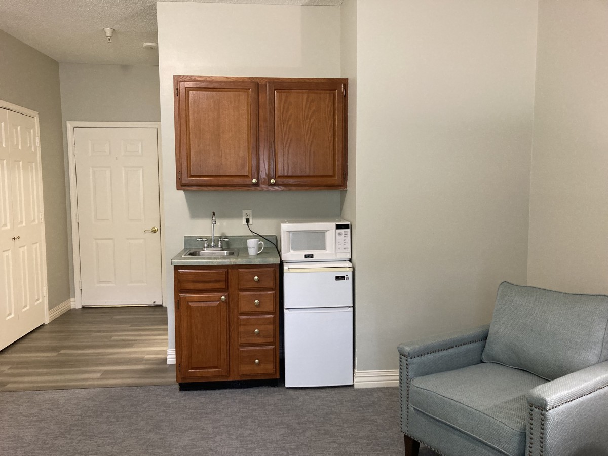 A compact kitchenette with wooden cabinets, a microwave, mini fridge, and sink next to a light blue armchair.