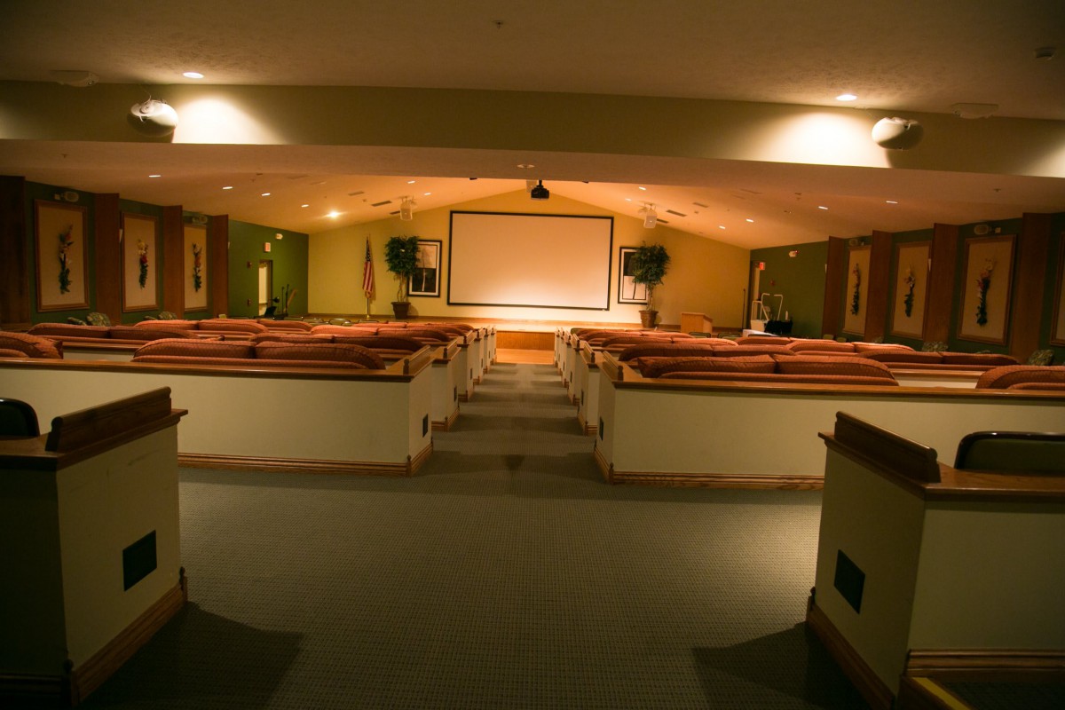 An empty auditorium with rows of wooden seats facing a large projector screen.