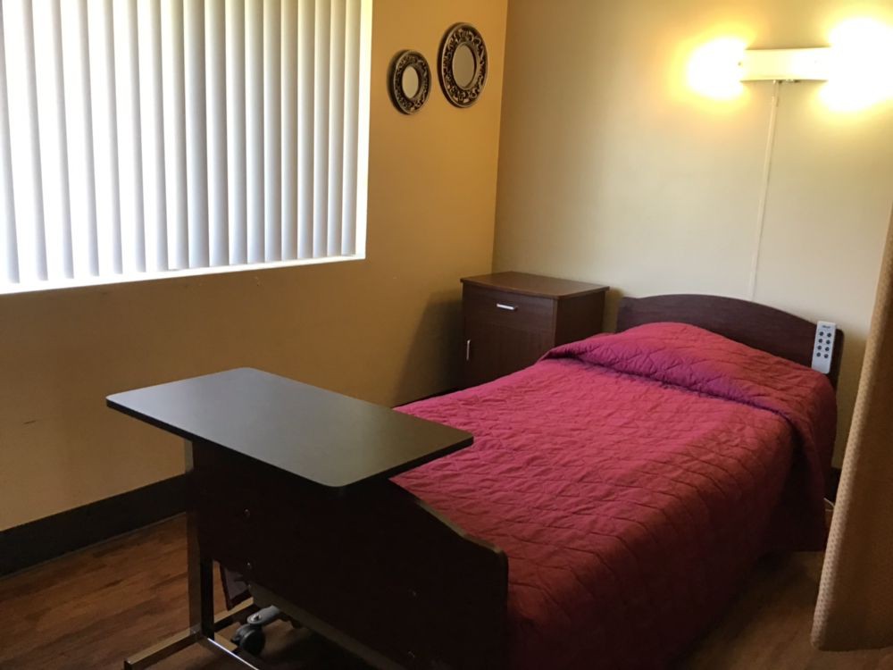 A minimalist bedroom with a single bed featuring a red bedspread and an overbed table, accompanied by wall decorations and window blinds.