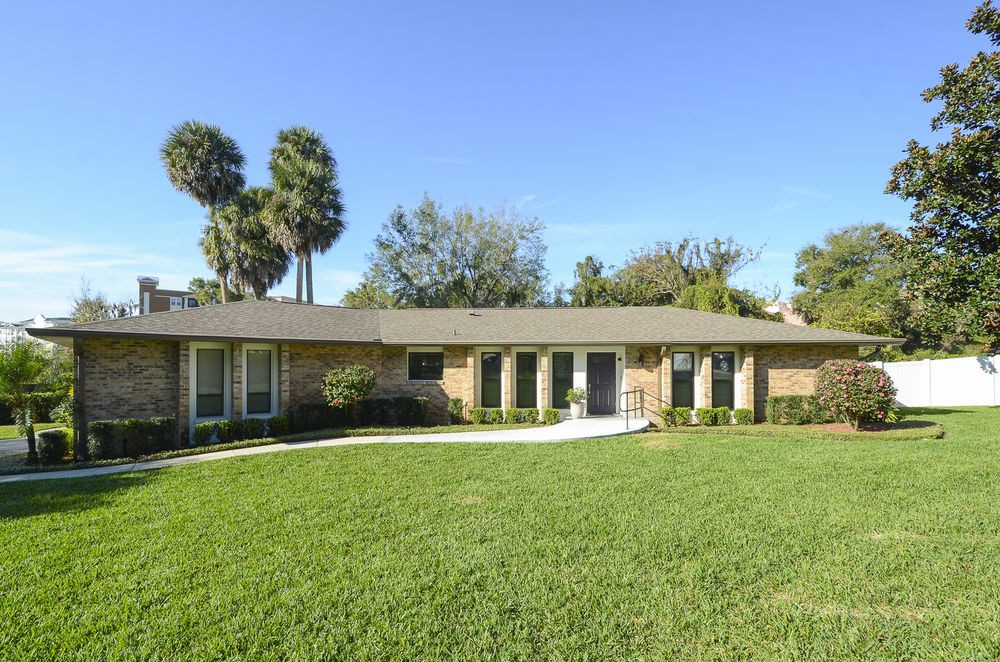 A one-story suburban house with a large lawn and palm trees in the background.