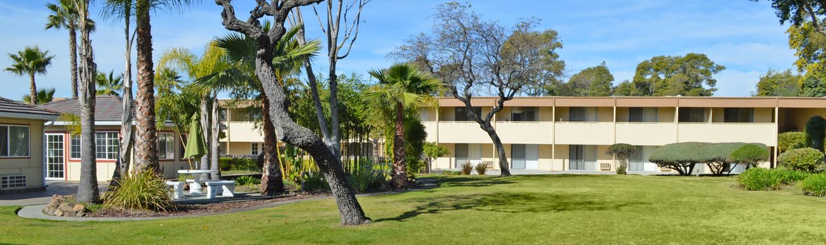 A two-story building surrounded by lush greenery and trees on a sunny day.