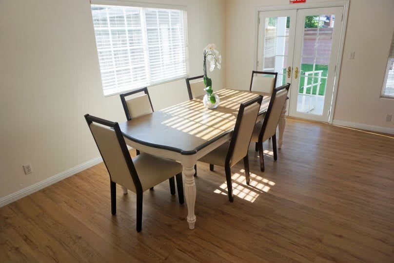 A bright dining room with a wooden table and six chairs, illuminated by sunlight streaming through the windows.