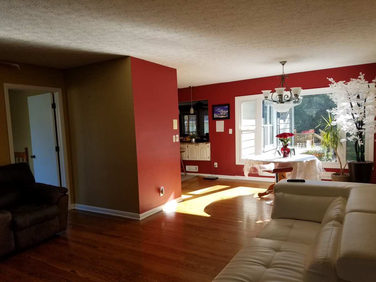A cozy living room with red walls and hardwood flooring, featuring a dining area and chandelier bathed in natural sunlight.