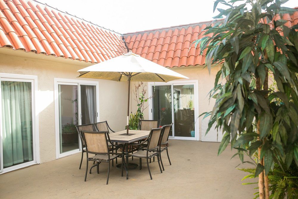 A cozy patio area with a table, chairs, and an umbrella, surrounded by plants and enclosed by sliding glass doors under a terracotta roof.