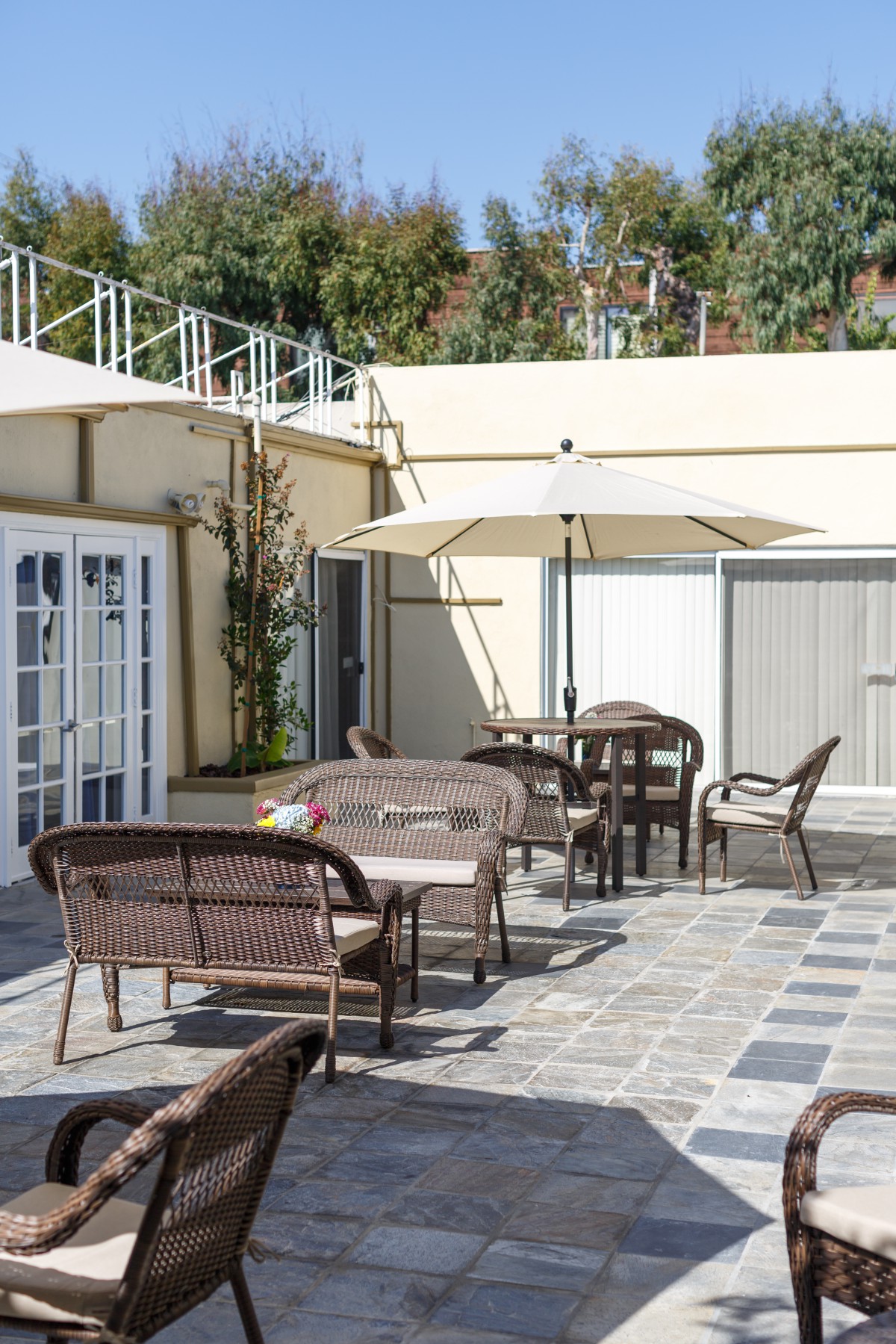 A sunny outdoor patio with wicker furniture, tables, chairs, and an umbrella.