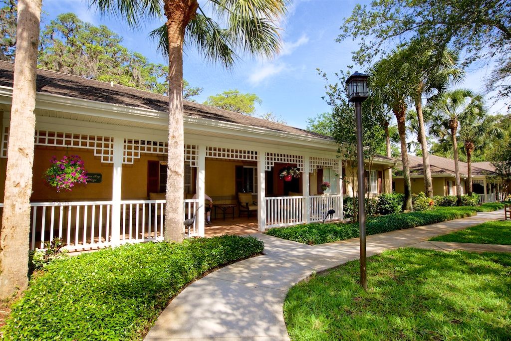A charming house with a welcoming porch, surrounded by lush greenery and palm trees, featuring a curved pathway leading to the entrance.