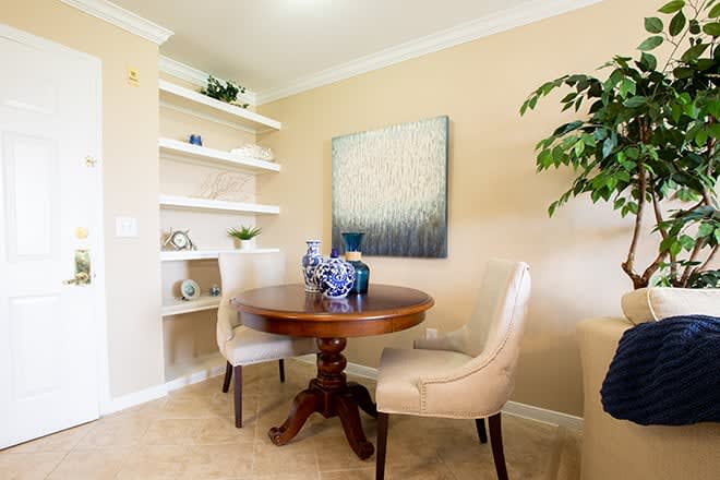 A cozy dining area with a round wooden table, upholstered chairs, and decorative elements on shelves.