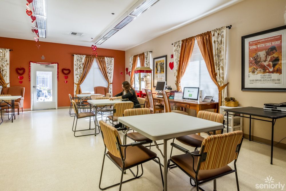 A cozy dining area with tables and chairs, decorated with curtains and wall hangings, featuring a computer station by the window.