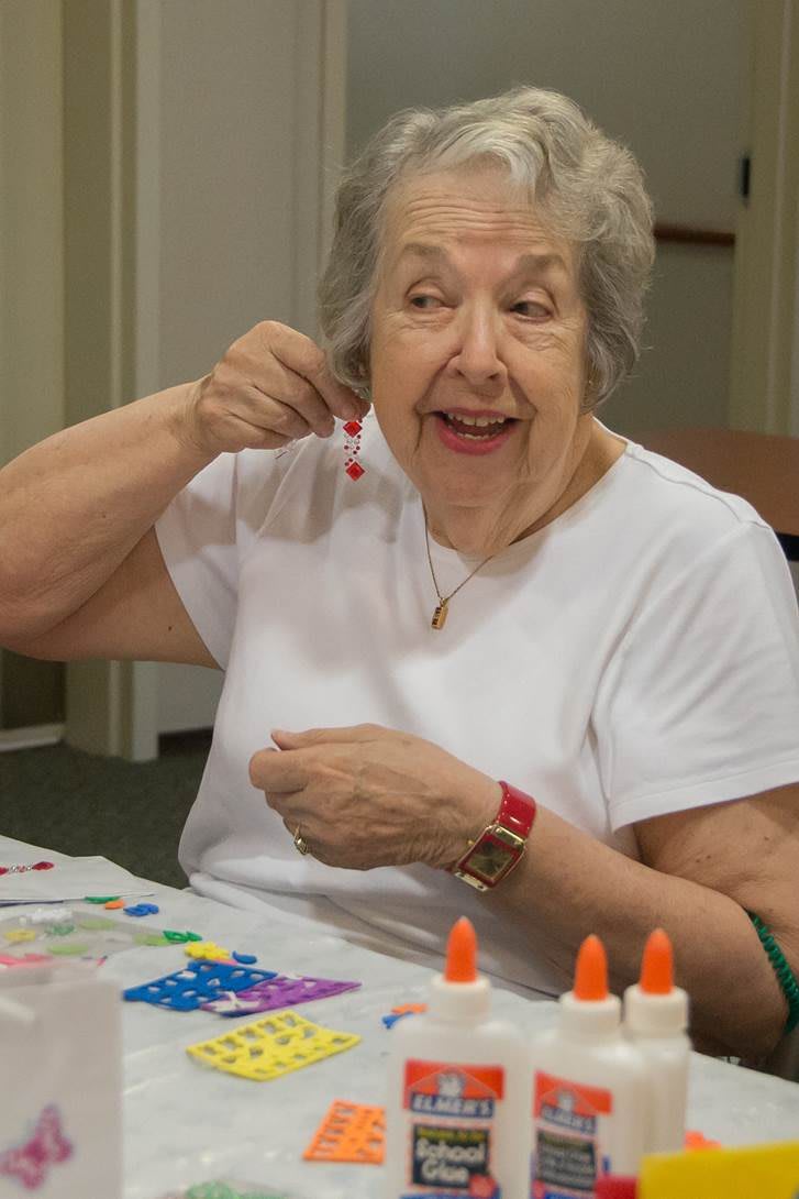 An elderly woman enjoying arts and crafts, smiling as she holds an earring near her ear.