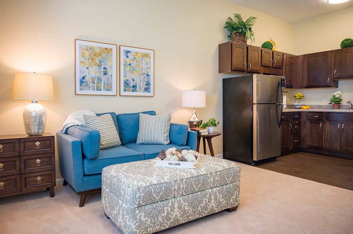 A cozy living room with a blue sofa, patterned ottoman, and decorative elements next to an open kitchen area.