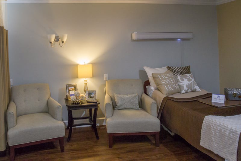 A cozy bedroom setting with two chairs, a side table, and a well-lit lamp beside a neatly made bed.