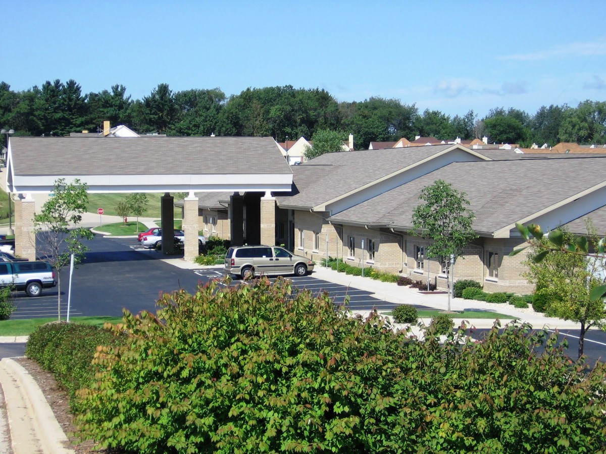 A view of a building with a parking lot and landscaping, featuring trees and bushes.