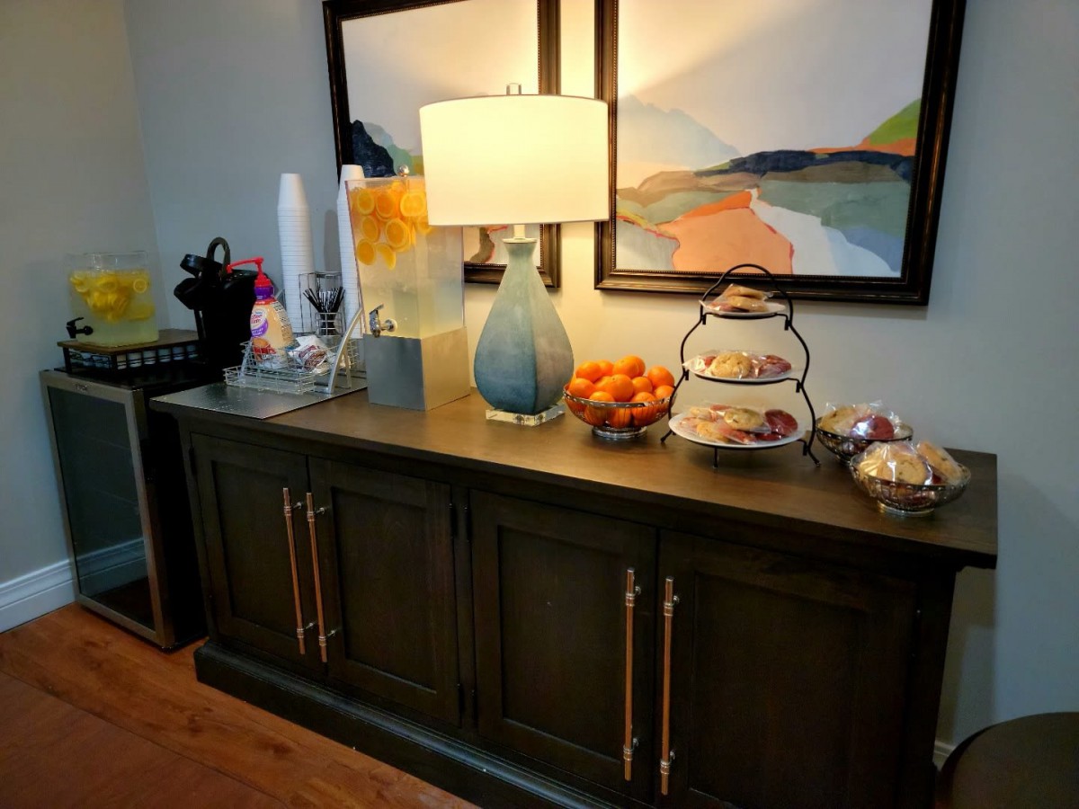 A stylish sideboard setup featuring a lamp, artwork, fruit bowl, and coffee station.
