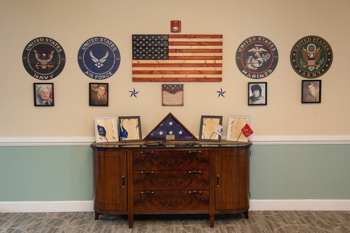 A wall display honoring military service, featuring an American flag, military seals, and framed photographs over a wooden cabinet.