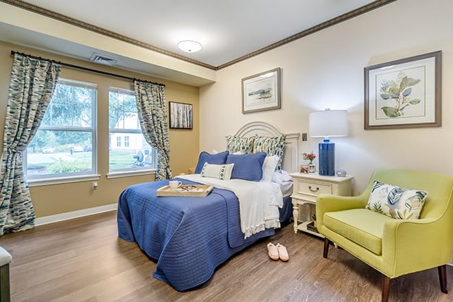 A cozy bedroom with a blue-themed bedspread, decorative pillows, and a green armchair next to a window with curtains.