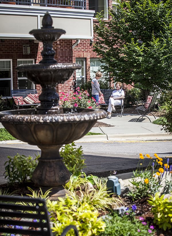 A tranquil outdoor setting featuring a tiered fountain, surrounded by lush landscaping and vibrant flowers, with benches in the background.