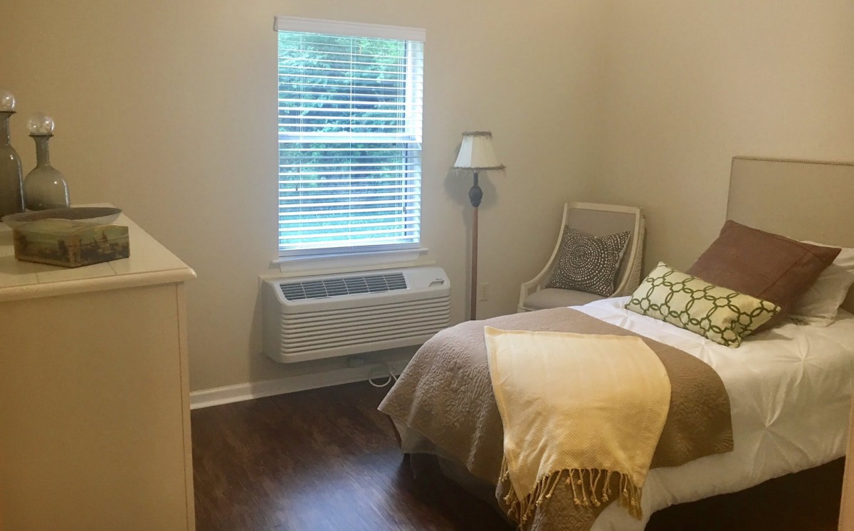 A cozy bedroom with a single bed, lamp, chair, and window air conditioner.