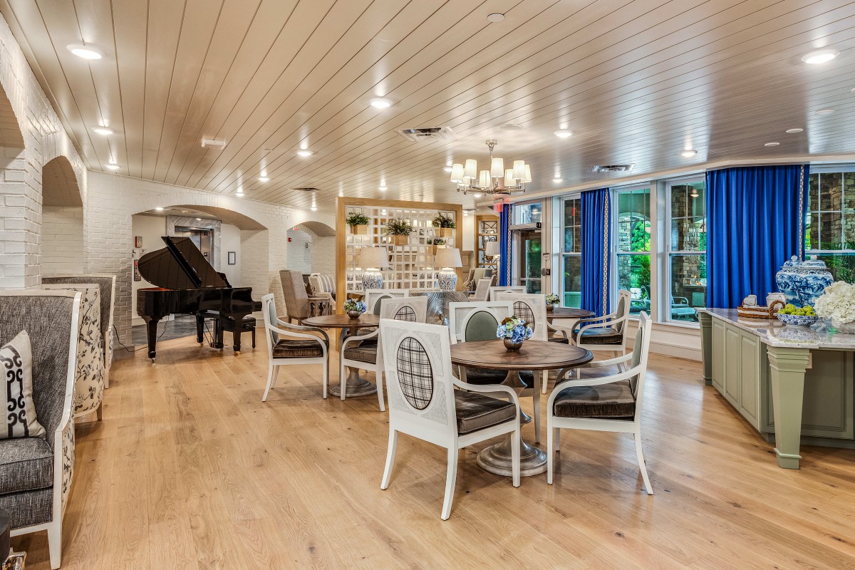 Elegant dining area with a piano and chandelier, featuring wood flooring and bright lighting.