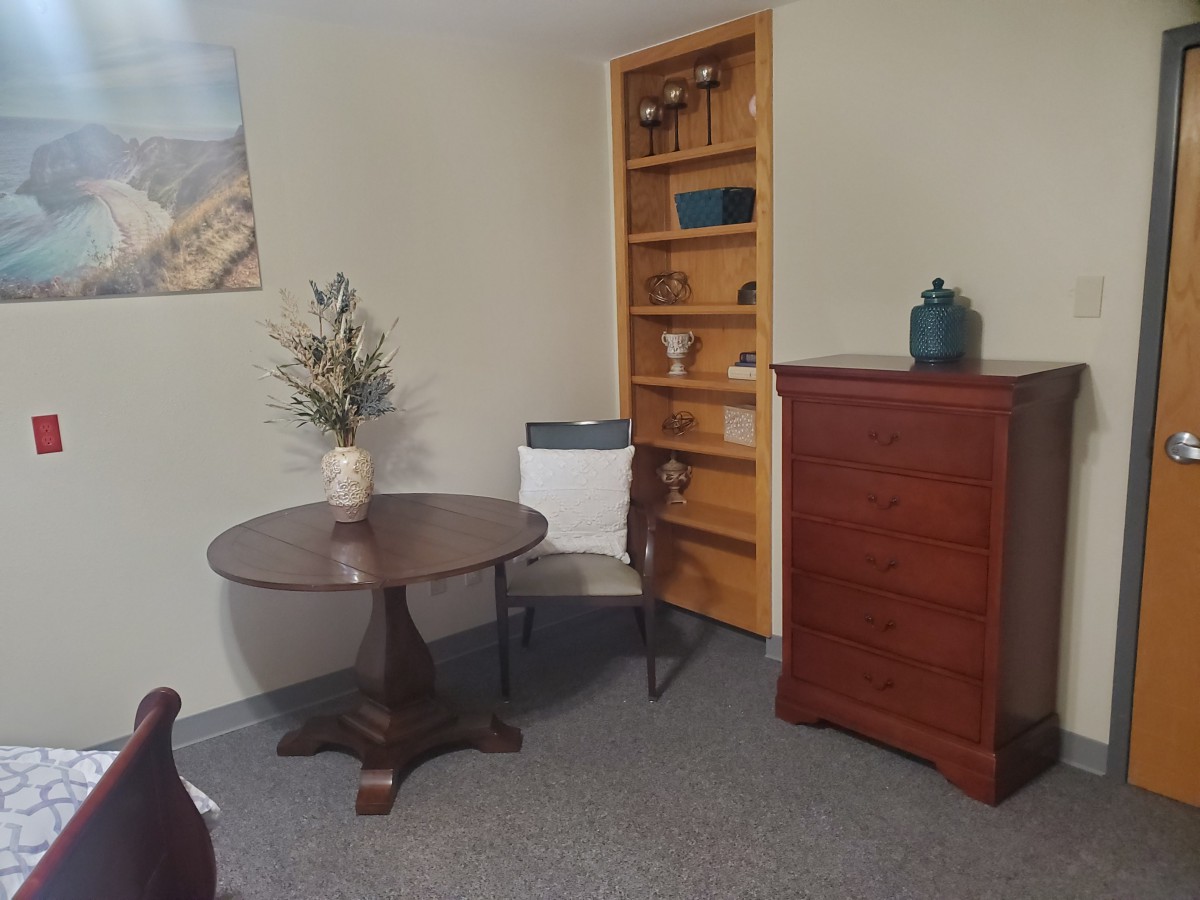 A cozy office corner with a round wooden table, a chair, and a tall bookshelf.