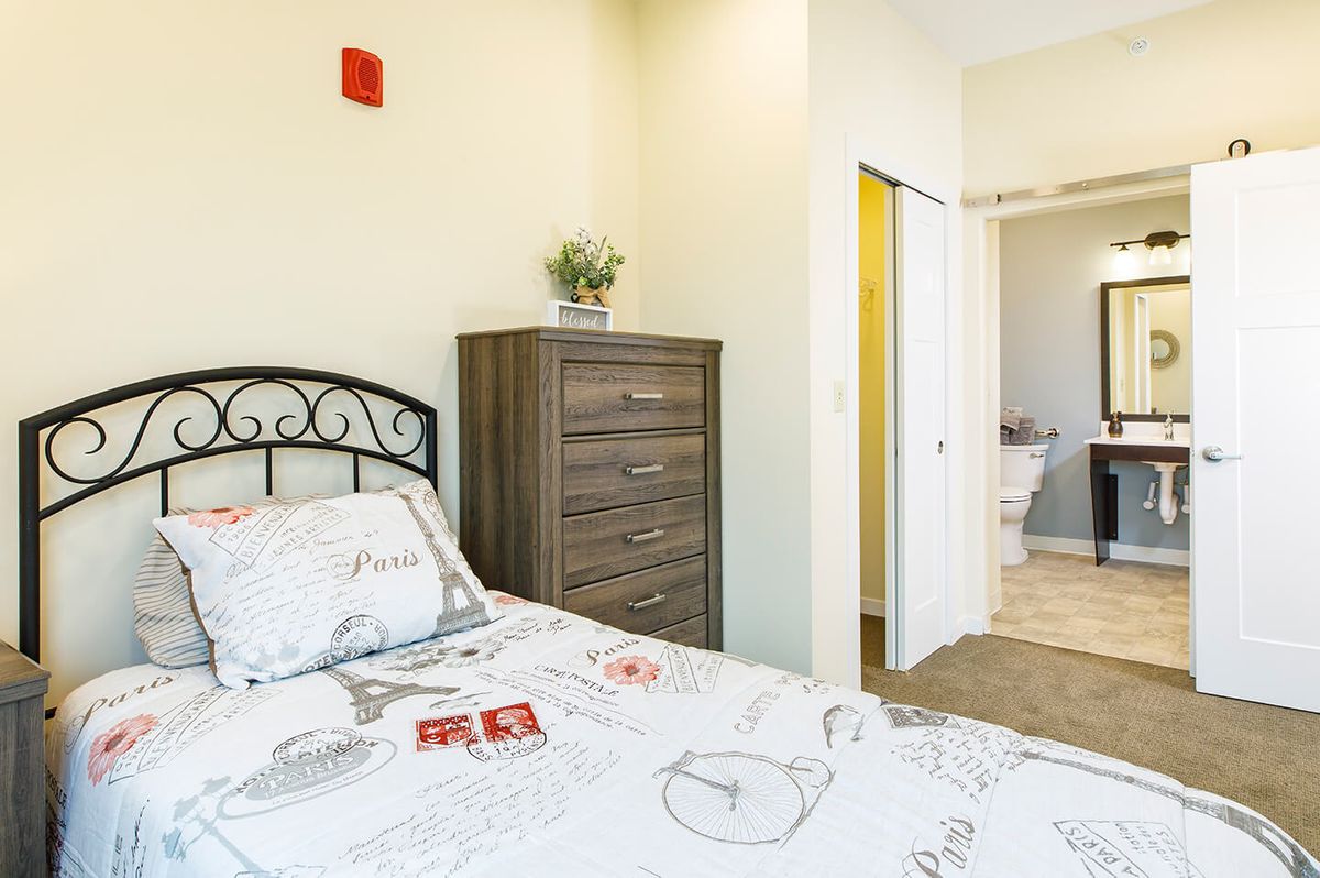 A cozy bedroom with a neatly made bed, a wooden dresser, and an open view into the bathroom.