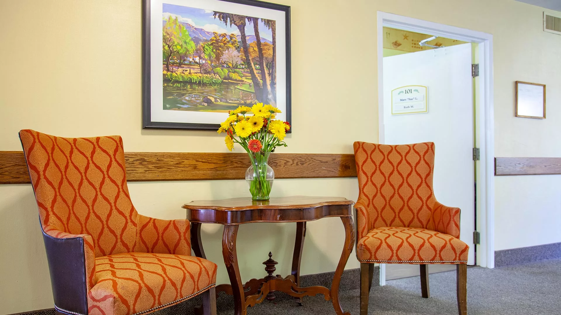 A cozy corner with two patterned orange chairs, a wooden table with a vase of yellow flowers, and a landscape painting on the wall.